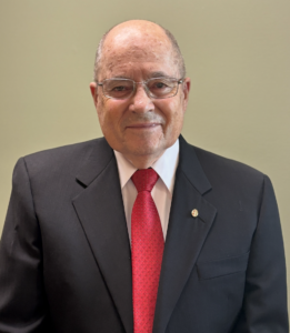 Headshot of Ed Gambs wearing a black suit against an olive green background